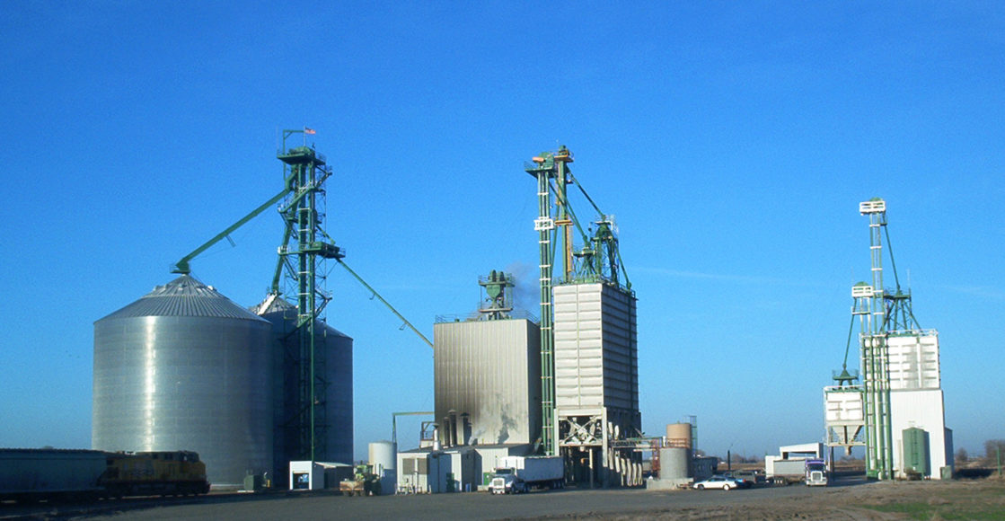 JDH Gooding mill with two grain bins, and two other large buildings for mixing and making feed ingredients. There is a bright blue sky and a train stopped in front of the bins.